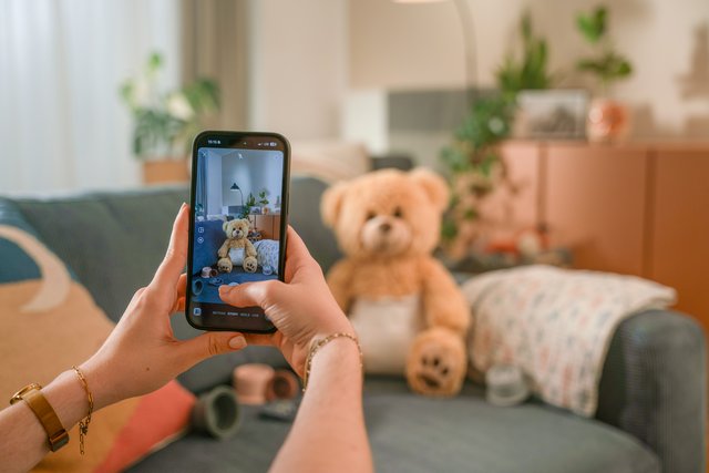 Man sieht zwei Hände die ein Mobiltelefon halten und ein Foto von einem Teddy Bär erstellen.