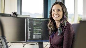 Mitarbeiterin lächelt in die Kamera. Im Hintergrund ist ihr Bildschirm mit Programmiercode drauf zu sehen.