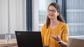 Mitarbeiterin sitzt vor einem Laptop und erklärt etwas mit Handgestiken. Weitere Mitarbeiterin hört zu. 