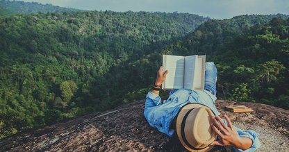Ein Mann liegt im grünen mit Blick auf den Wald und lest ein Buch.