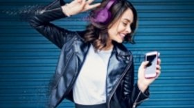 Frau die mit Kopförern Musik horcht, in einer Hand ein Mobiltelefon, mit der anderen Hand V-Zeichen macht