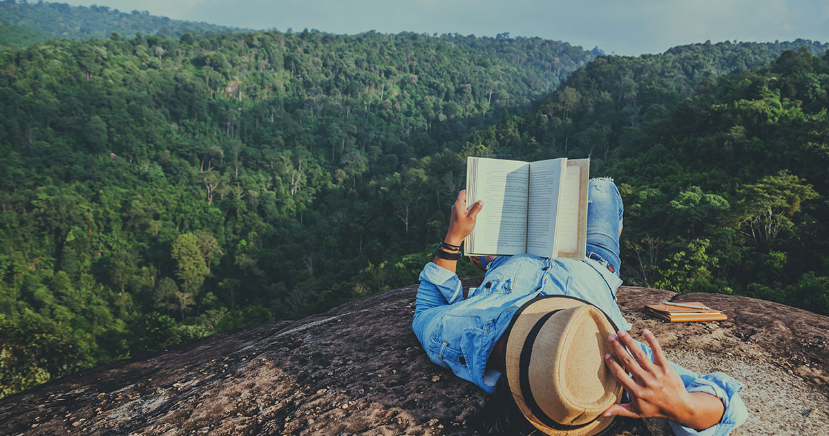 Ein Mann liegt im grünen mit Blick auf den Wald und lest ein Buch.