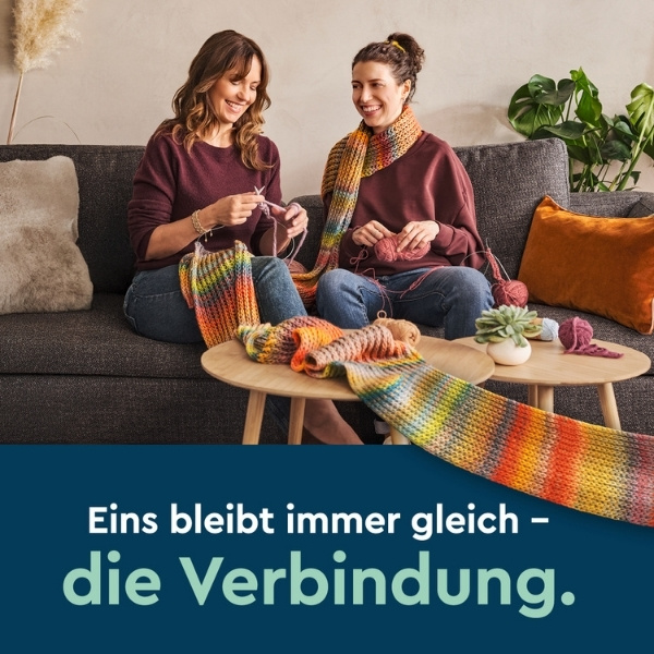 Mutter & Tochter auf der Couch beim Stricken eines extrem langen, bunten Schals mit dunkelblauem Hintergrund, Text: Eins bleibt immer gleich – die Verbindung.