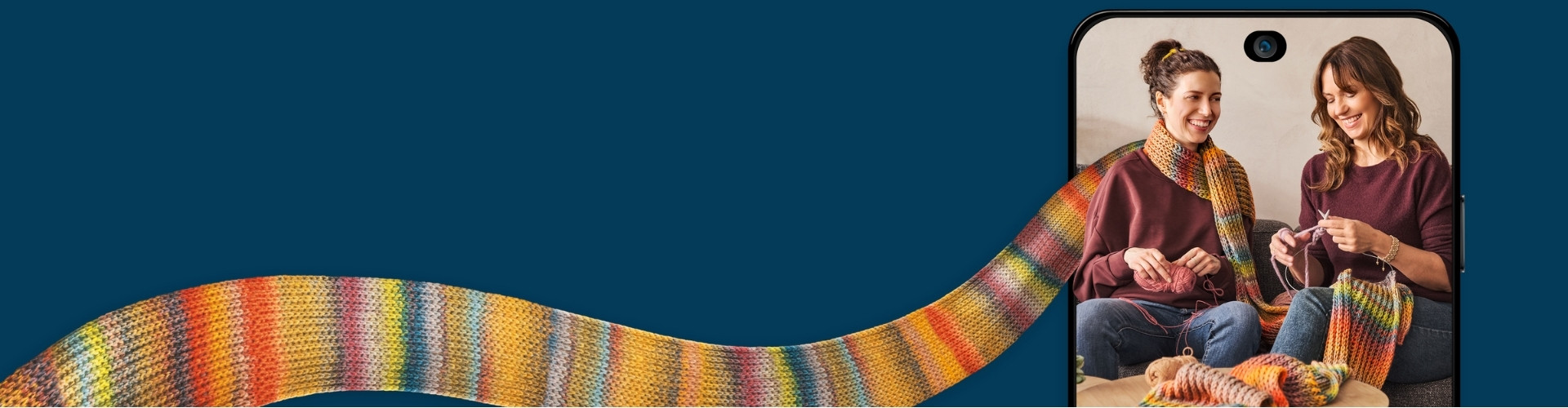 Mutter & Tochter auf der Couch beim Stricken eines extrem langen, bunten Schals mit dunkelblauem Hintergrund