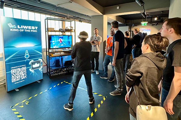 LIWEST Stand mit Spiel King of the Ping VR. Ein junger Mann mit VR-Brille aufgesetzt, spielt das Spiel. Dahinter Publikum. LIWEST Stand mit Spiel King of the Ping VR. Ein junger Mann mit VR-Brille aufgesetzt, spielt das Spiel. Dahinter Publikum.