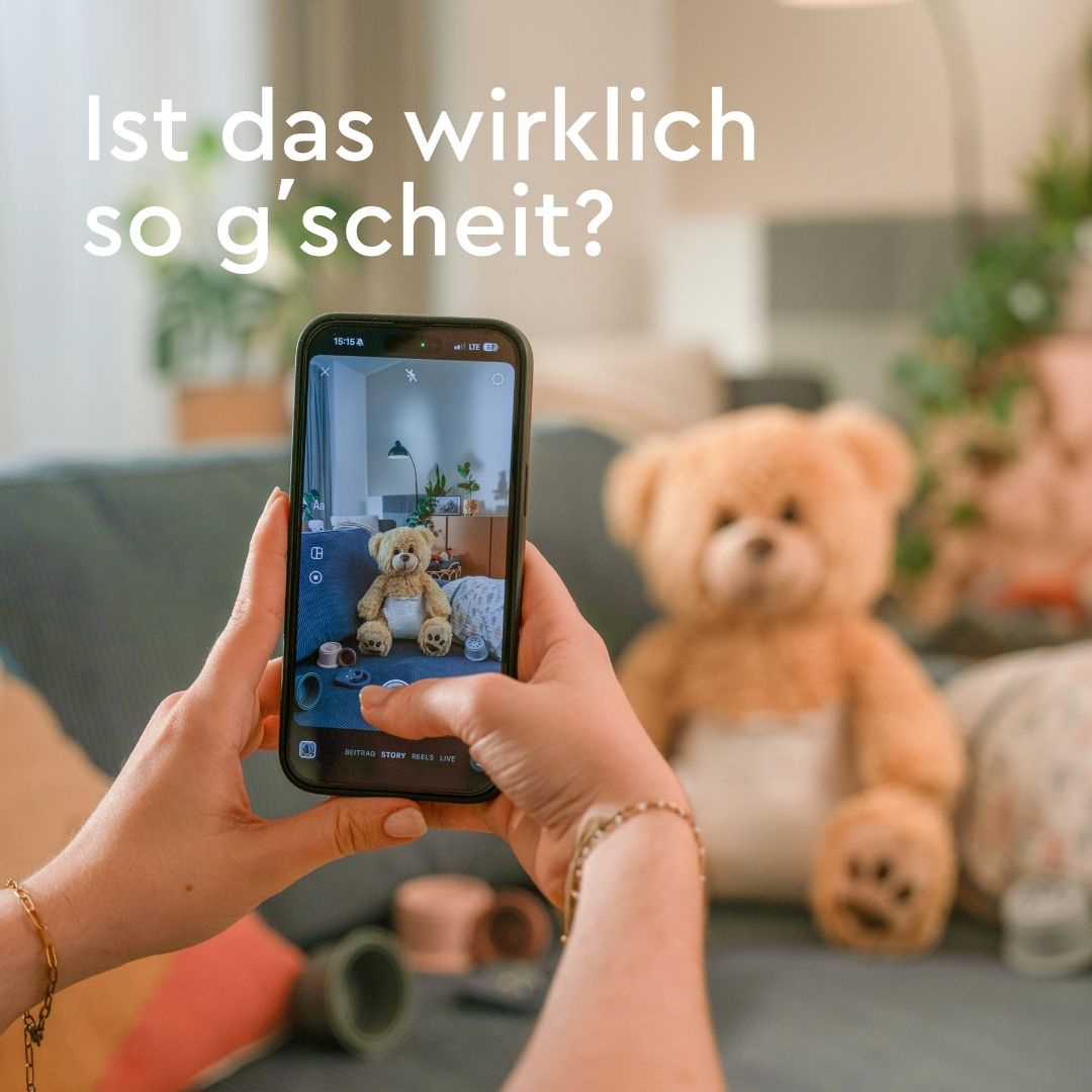 Text "Ist das wirklich so g'scheit?" mit Hand/Handy das Teddy auf Couch fotografiert