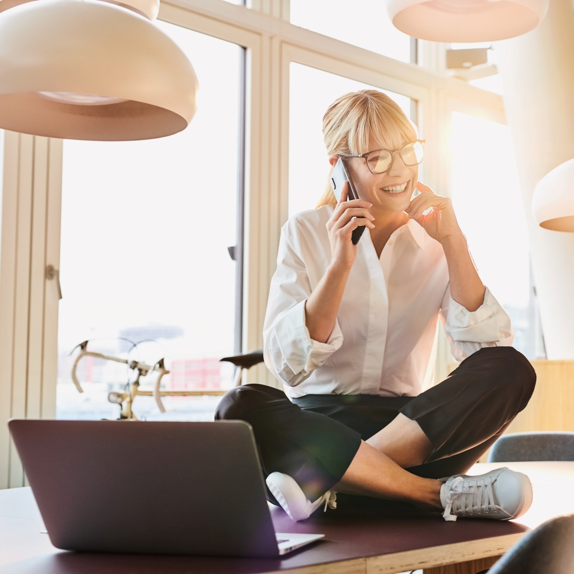 Blonde Frau mit weißer Bluse sitzt im Schneidersitz vor dem Laptop und telefoniert mit Smartphone